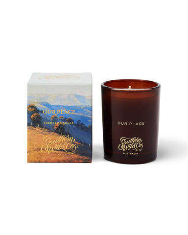 An amber glass candle alongside its kraft box, which features an illustration of a bush landscape.
