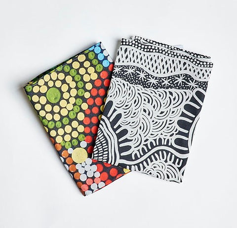 Two folded colorful Indigenous artwork tea towels, one with a dot pattern in vibrant colors and another with a black and white pattern.
