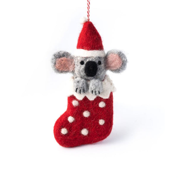 A felt koala ornament wearing a red Santa hat and sitting in a red stocking with white polka dots, hanging from a red and white string on a white background.
