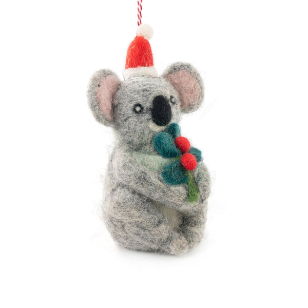 A felted koala ornament wearing a Santa hat and holding green holly with red berries, hanging from a red and white string against a white background.