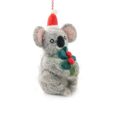 A felted koala ornament wearing a Santa hat and holding green holly with red berries, hanging from a red and white string against a white background.