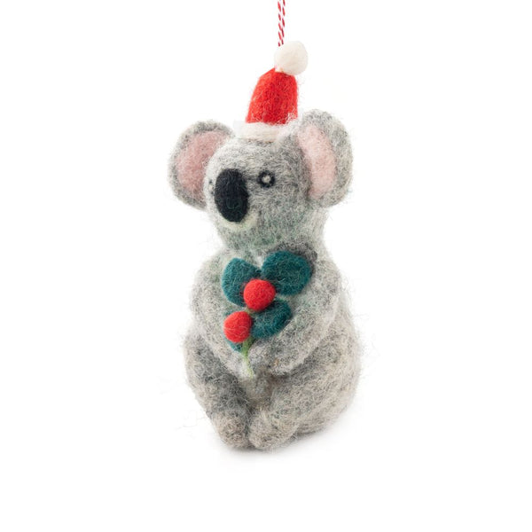 A felted koala ornament wearing a Santa hat and holding green holly with red berries, hanging from a red and white string against a white background.