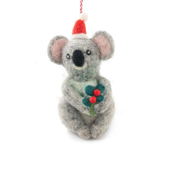 A felted koala ornament wearing a Santa hat and holding green holly with red berries, hanging from a red and white string against a white background.