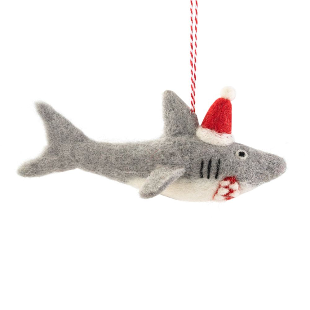 A felted shark Christmas ornament, wearing a red Santa hat, hanging from a red-and-white striped string against a white background.
