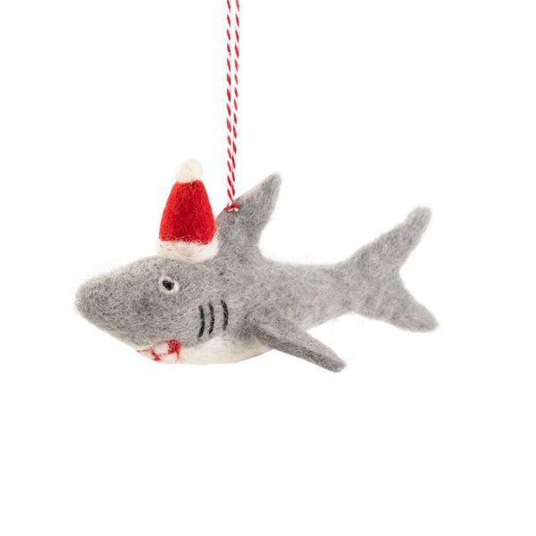 A felted shark Christmas ornament, wearing a red Santa hat, hanging from a red-and-white striped string against a white background.
