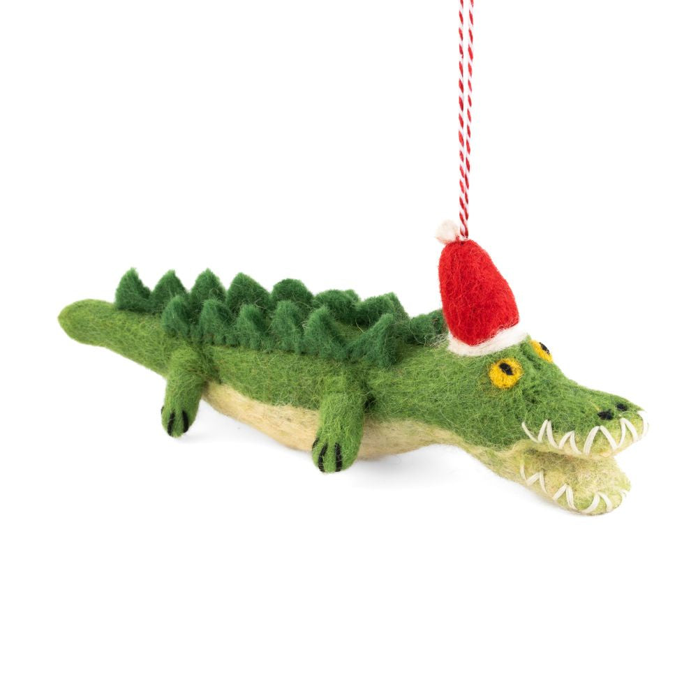 A felted crocodile Christmas ornament wearing a red Santa hat, hanging from a red and white string against a white background.