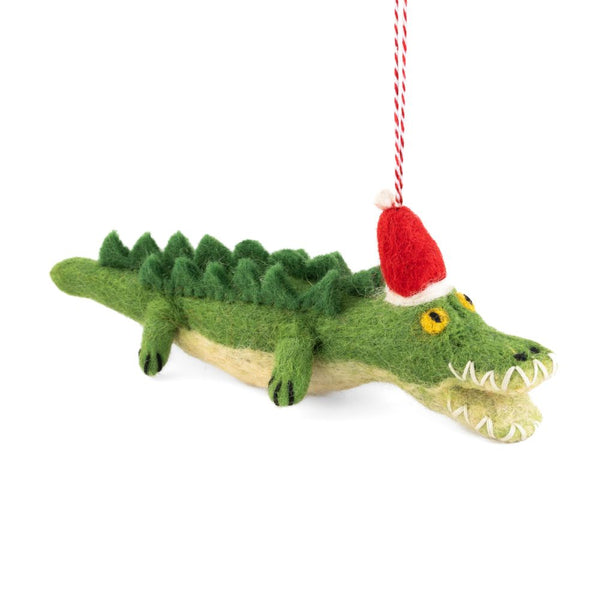 A felted crocodile Christmas ornament wearing a red Santa hat, hanging from a red and white string against a white background.