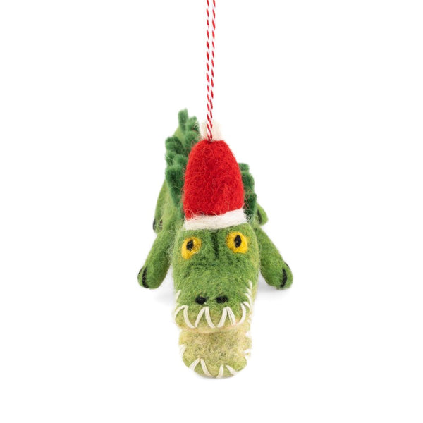 A felted crocodile Christmas ornament wearing a red Santa hat, hanging from a red and white string against a white background.