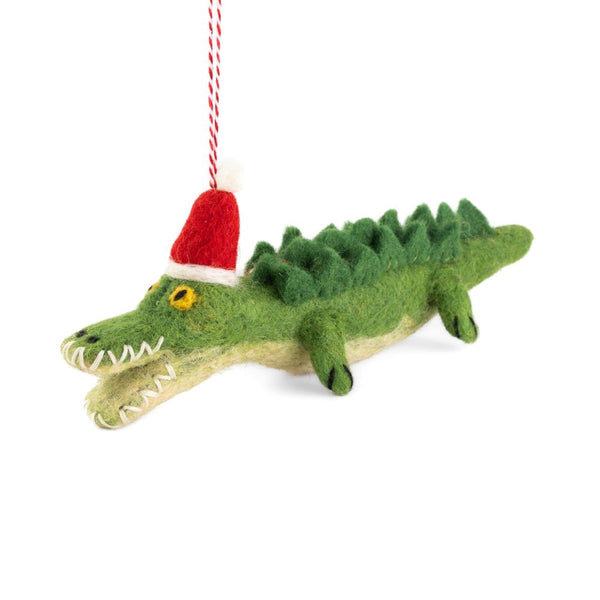A felted crocodile Christmas ornament wearing a red Santa hat, hanging from a red and white string against a white background.