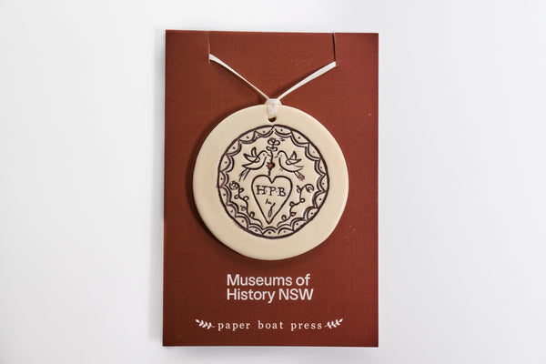 Ceramic convicr Love Token ornament on a backing card with 'Museums of History NSW' and 'paper boat press' text.