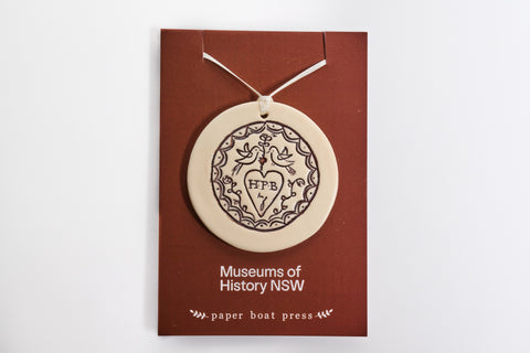 Ceramic convicr Love Token ornament on a backing card with 'Museums of History NSW' and 'paper boat press' text.