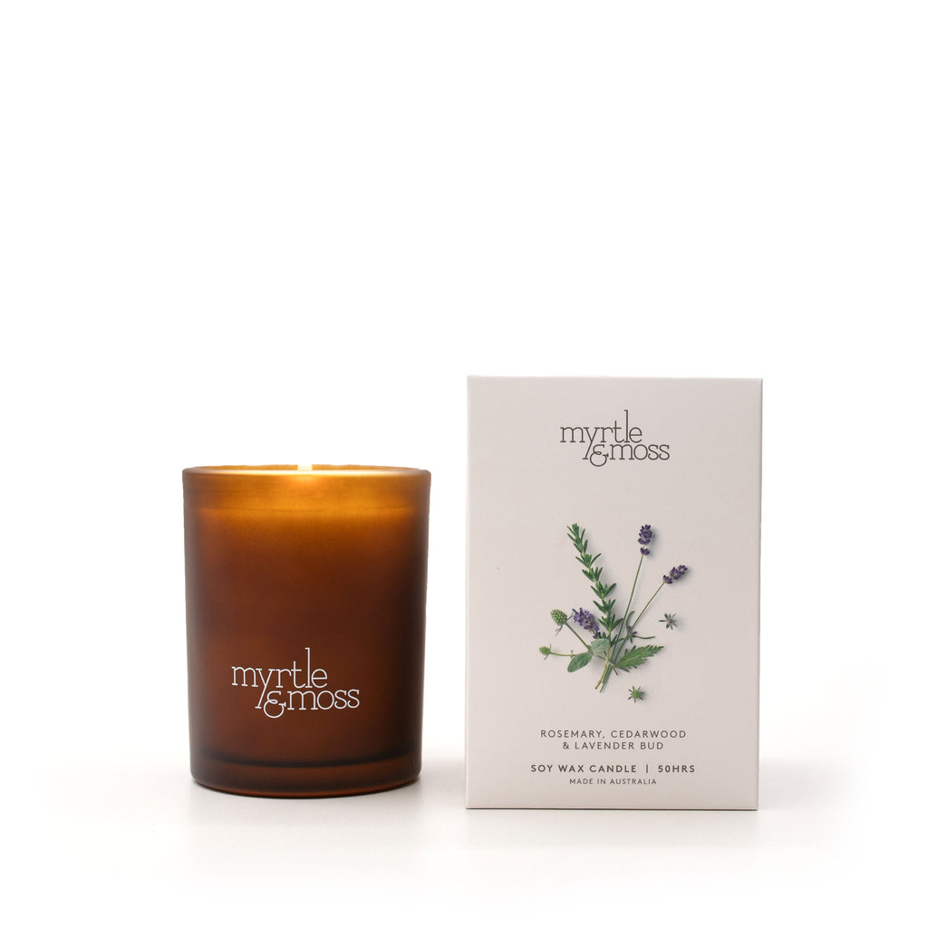 An amber glass soy wax candle with a lit flame stands next to its white packaging box on a clean white background. The box features a botanical illustration of rosemary and lavender with the brand name 'myrtle & moss'.