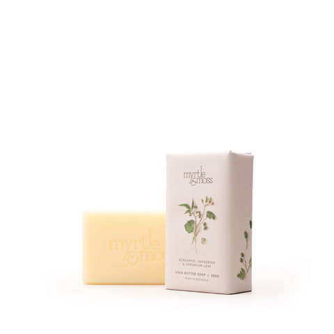 A pale yellow bar of Myrtle & Moss Shea & Butter Soap stands next to its cream-colored rectangular packaging box, which features a botanical illustration of a green leaf sprig.