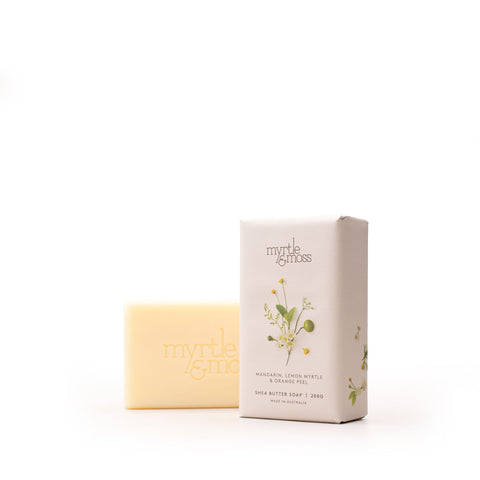 A pale yellow bar of Myrtle & Moss Shea Butter Soap stands next to its cream-colored packaging box, which features a small botanical illustration of lemon myrtle, on a seamless white background.