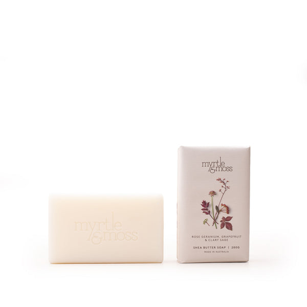 A bar of Myrtle & Moss unscented grapefruit and clary sage milk butter soap stands next to its paper packaging box on a seamless white background.