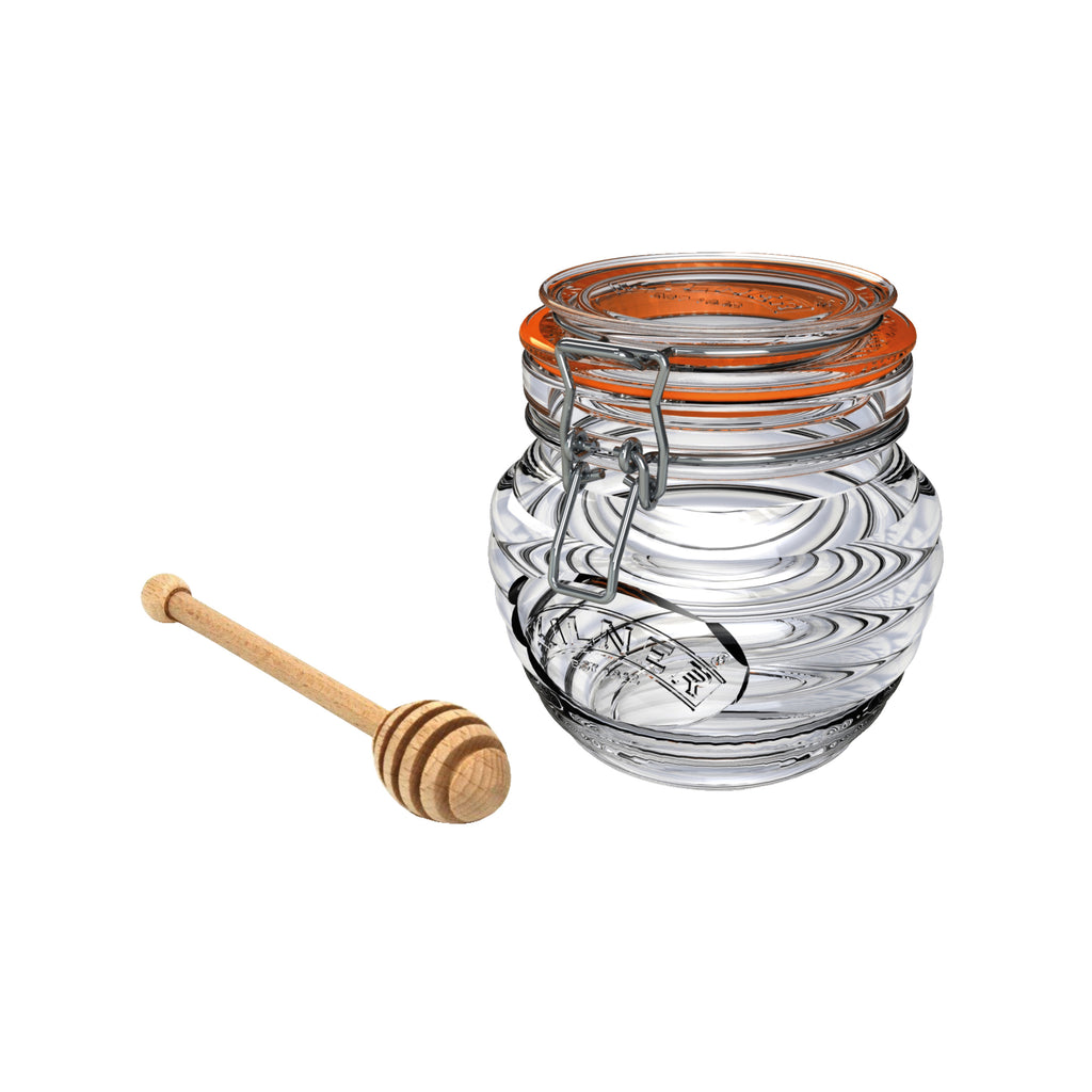 A clear glass honey pot with a lid with metal hinge and clasp, and a beechwood honey dipper next to it.