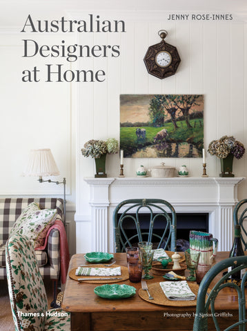 The cover of the book 'Australian Designers at Home' by Jenny Rose-Innes, featuring a living room with a white fireplace, wooden table, and green chairs, topped with a painting and a decorative clock.