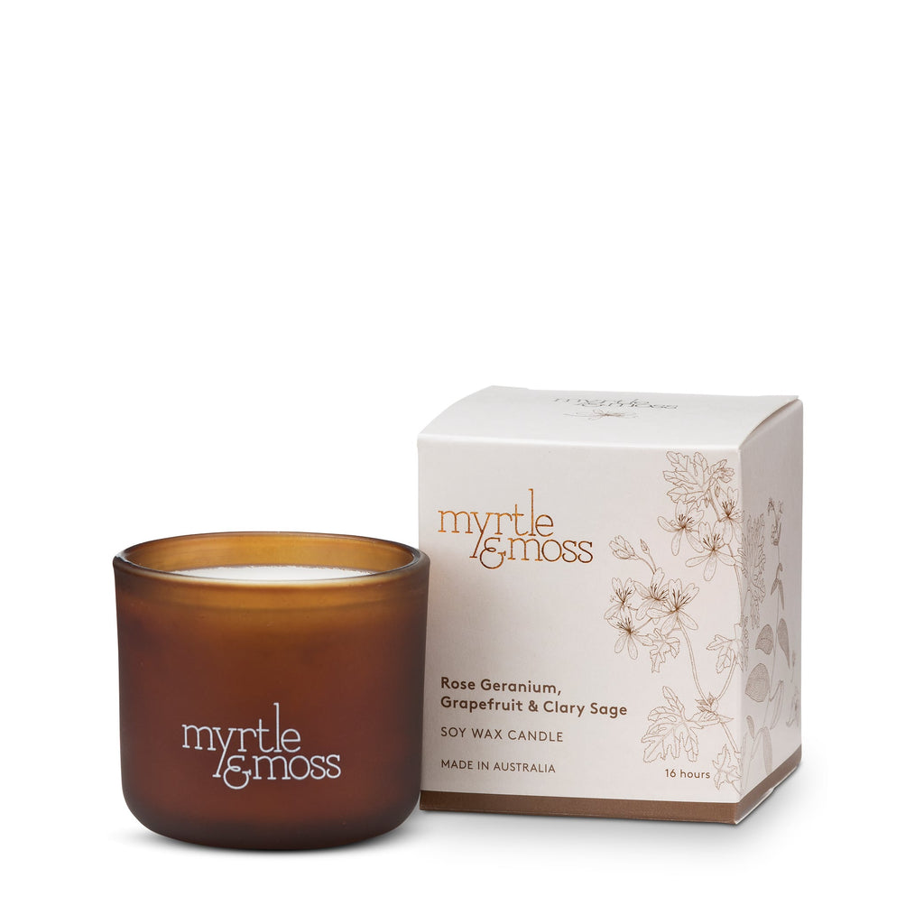 A small amber-colored soy wax candle next to its packaging box which has floral designs and the brand 'Myrtle & Moss' printed on it.