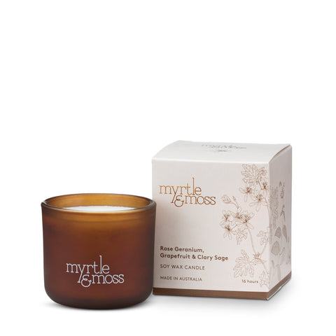 A small amber-colored soy wax candle next to its packaging box which has floral designs and the brand 'Myrtle & Moss' printed on it.