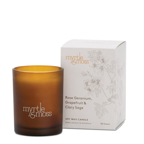 A soy wax candle housed in amber glass with rose geranium, grapefruit, and clary sage fragrance, alongside its packaging box with floral designs. The candle is labeled with 'Myrtle & Moss' branding.