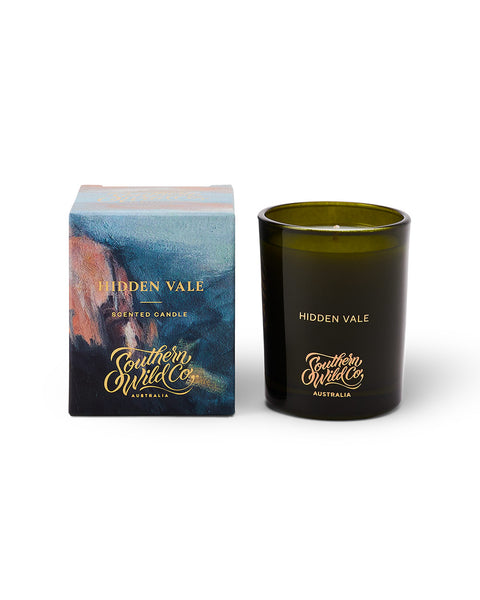 A dark green glass candle next to its kraft box with a landscape design and the text 'Southern Wild Co.'.