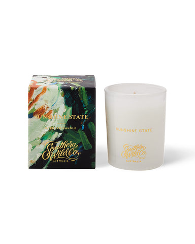 A white candle with a gold text, next to its packaging box featuring an artistic design with green, gold, and red colours.